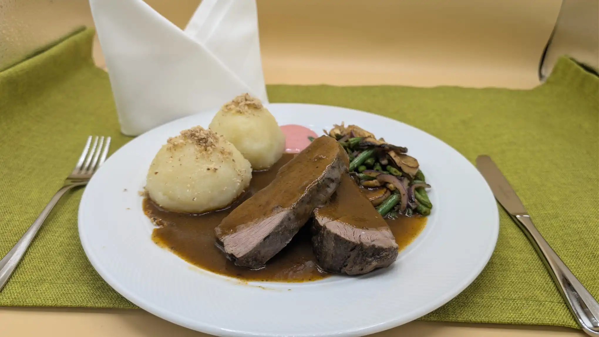 Zwei Scheiben Braten mit brauner Soße, zwei Knödel und Gemüse aus grünen Bohnen, Pilzen und Zwiebeln auf einem weißen Teller.